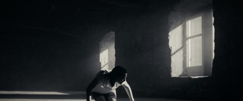 A woman crouches on the floor of a large dark concrete room with two lit windows.
