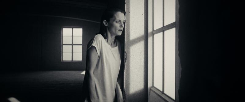 A woman leans against concrete beside a tall bright window, face half-lit.