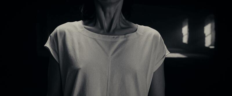 Torso and neck of a woman in a pale t-shirt, face out of frame, windows glowing behind.